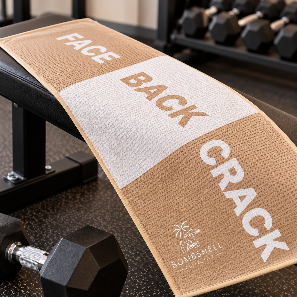Bombshell Collective Face Back Crack gym towel in sand displayed on a gym bench, featuring a bold three-section design.