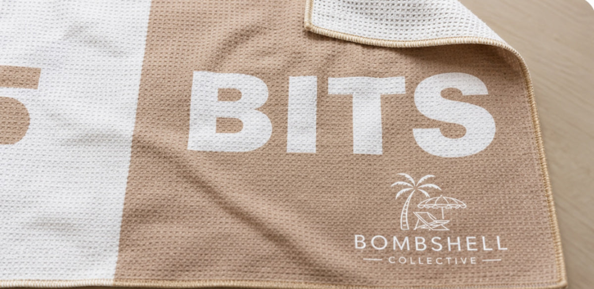 Close-up of the Face Bum Bits beach towel in sand, showing textured sand-free waffle microfibre fabric, folded corner and Bombshell Collective branding.